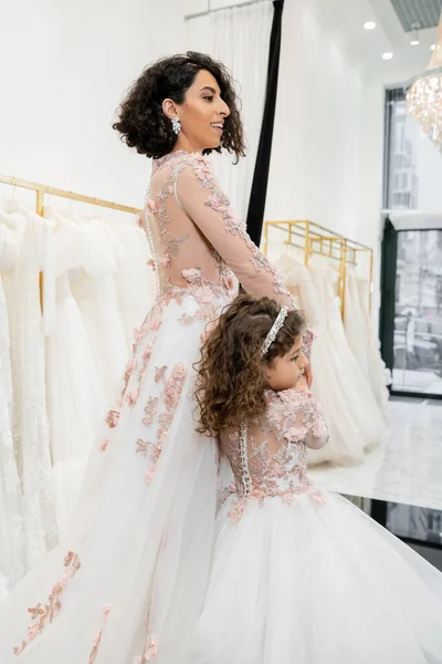 Cute middle eastern kid in floral attire holding hands with happy woman and walking in floral dresses near blurred wedding gown inside of bridal salon, shopping, bride-to-be, mother and daughter — Stock Photo