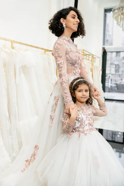 Chica de Oriente Medio en traje floral cogido de la mano con la madre alegre de pie en vestido de novia cerca borrosa vestido blanco dentro de lujoso salón de novia, compras, novia-a-ser, vinculación - foto de stock