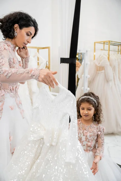 Brunette mariée du Moyen-Orient en robe de mariée florale aidant à choisir la robe pour sa petite fille dans le salon de mariée autour des tissus de tulle blanc, processus de préparation, ensemble — Photo de stock