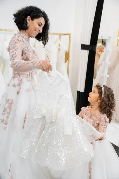 Glückliche und brünette mittelöstliche Braut in floralem Hochzeitskleid, die hilft, Kleid für ihre kleine Tochter auszuwählen, im Brautsalon um weiße Tüll-Stoffe, Vorbereitungsprozess, Zweisamkeit — Stockfoto