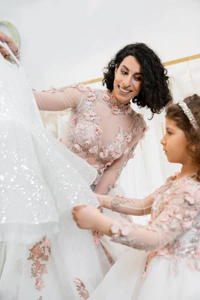 Charmante et brune femme du Moyen-Orient en robe de mariée fleurie aidant à choisir la robe pour sa petite fille dans le salon nuptial autour de tissus en tulle blanc, processus de préparation, convivialité — Photo de stock