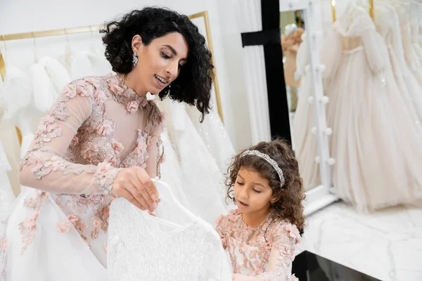 Brunette mariée du Moyen-Orient en robe de mariée florale aidant à choisir la robe pour sa jolie petite fille dans la boutique nuptiale autour de tissus en tulle blanc, processus de préparation, ensemble — Photo de stock