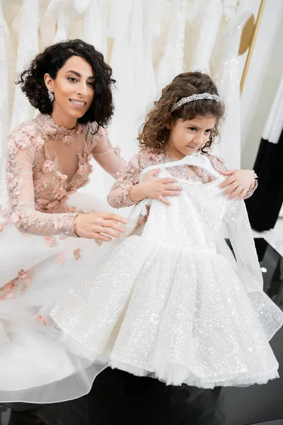 Happy and brunette middle eastern bride in floral wedding gown helping to choose dress for her little daughter in bridal salon around white tulle fabrics, process of preparation — Stock Photo