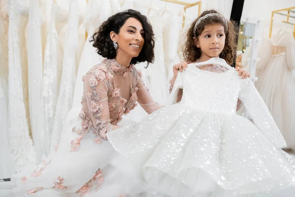 Cheerful and brunette middle eastern bride in floral wedding gown looking away near her little daughter with cute dress in bridal salon around white tulle fabrics, process of preparation — Stock Photo