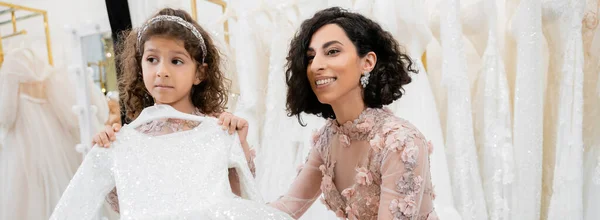 Bezaubernde und brünette mittelöstliche Braut in floralem Hochzeitskleid, die hilft, Kleid für ihre kleine Tochter im Brautsalon um weiße Tüll-Stoffe zu wählen, Vorbereitungsprozess, Banner — Stockfoto