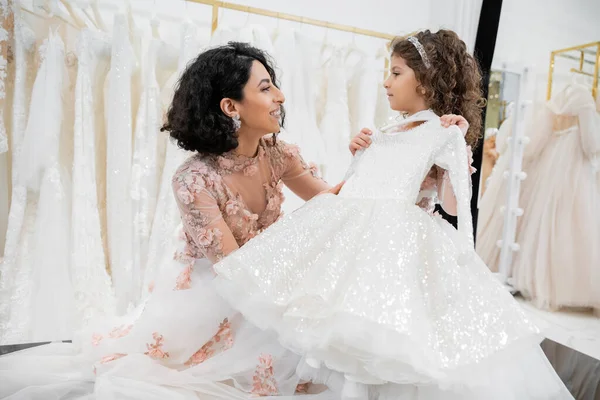 Happy and brunette middle eastern woman in floral wedding gown helping to choose white dress for her little daughter in bridal salon around tulle fabrics, process of preparation — Stock Photo
