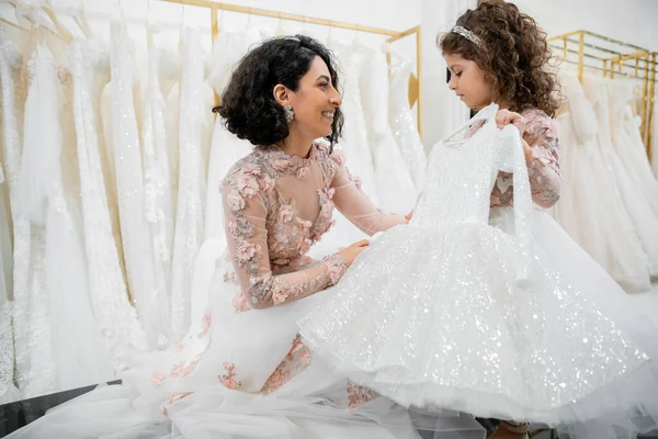 Blissful and brunette middle eastern bride in floral wedding gown helping to choose dress for her cute little daughter in bridal salon around white tulle fabrics, process of preparation — Stock Photo