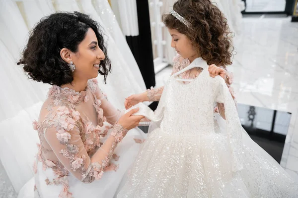 Happy and brunette middle eastern bride in floral gown helping her little daughter to choose dress in bridal salon around white tulle fabrics, process of preparation for wedding — Stock Photo