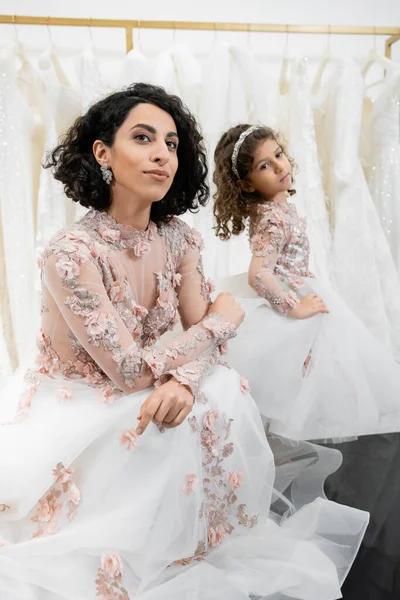 Moment spécial, mariée brune du Moyen-Orient en robe de mariée florale assise à côté de sa petite fille dans un salon nuptial autour de tissus en tulle blanc, shopping nuptial, convivialité — Photo de stock