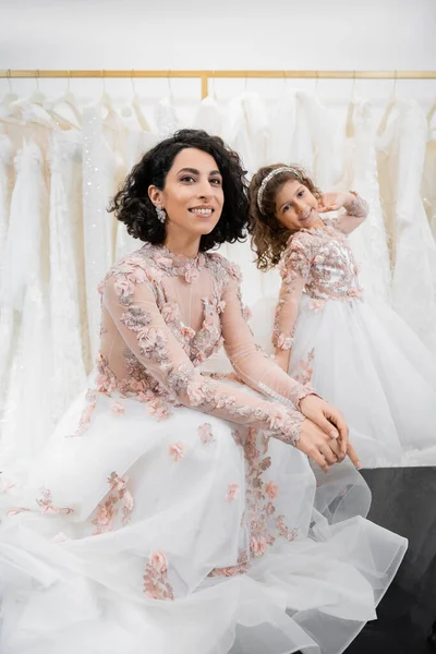 Momento especial, encantadora y feliz novia de Oriente Medio en vestido de novia floral sentado junto a su hija pequeña en el salón de novia alrededor de telas de tul blanco, compras nupciales, juntos - foto de stock