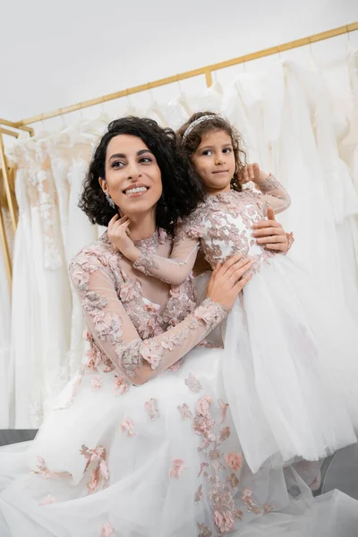 Momento especial, encantadora novia de Oriente Medio en vestido de novia floral abrazando a su hija pequeña en el salón de novia alrededor de telas de tul blanco, compras nupciales, juntos - foto de stock