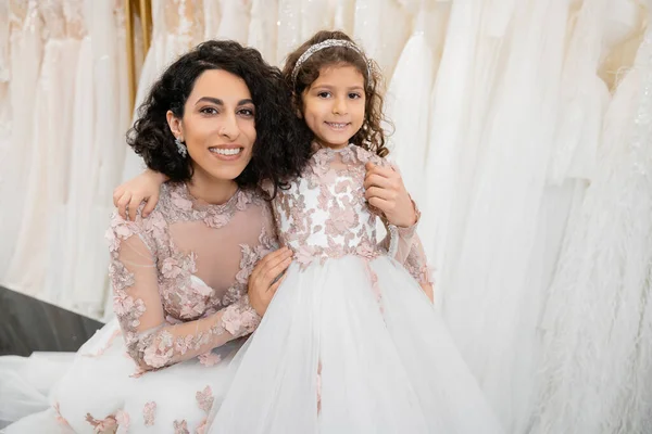 Moment spécial, heureuse femme du Moyen-Orient en robe de mariée florale assise et embrassant sa petite fille dans un salon nuptial autour de tissus en tulle blanc, shopping nuptial, convivialité, future mariée — Photo de stock