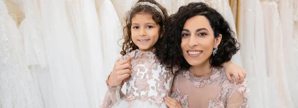 Momento especial, alegre novia de Oriente Medio en vestido de novia floral sentado y abrazando a su hija pequeña en el salón de novia alrededor de telas de tul blanco, compras nupciales, unión, bandera - foto de stock