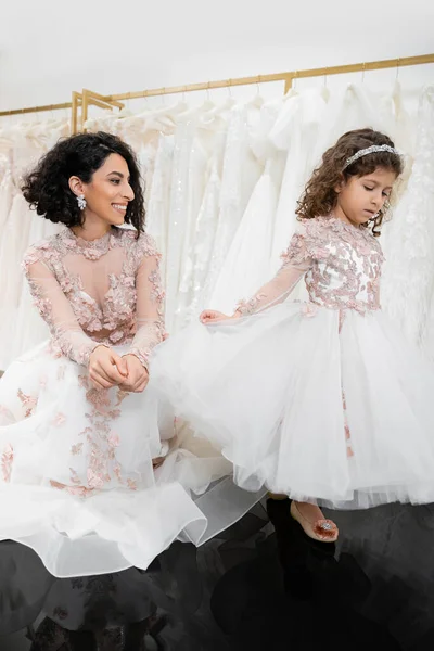 Momento especial, alegre noiva do Oriente Médio em vestido de casamento floral sentado e olhando para a filhinha no salão de casamento posando em torno de tecidos de tule branco, compras nupciais, união — Fotografia de Stock