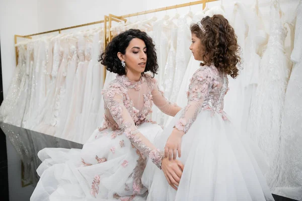 Особенный момент, брюнетка Ближнего Востока невеста в цветочном свадебном платье регулируя юбку наряд ее маленькой дочери в свадебном салоне вокруг белых тканей тюля, свадебные покупки, единение — стоковое фото