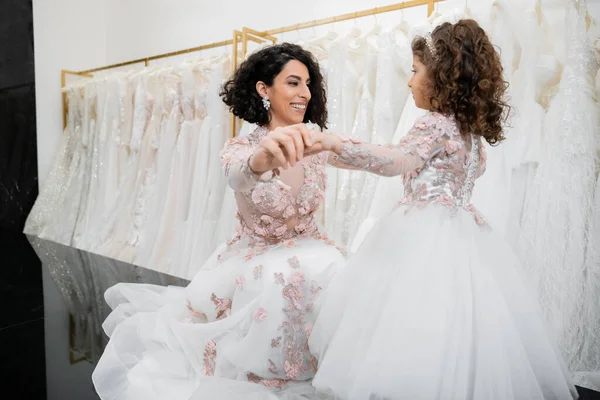 Moment spécial, heureuse mariée du Moyen-Orient en robe de mariée florale assise et tenant la main avec sa petite fille dans le salon nuptial autour de tissus en tulle blanc, shopping nuptial, convivialité — Photo de stock