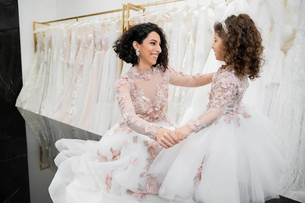 Moment spécial, heureuse mariée du Moyen-Orient en robe de mariée assise et tenant la main avec sa petite fille dans le salon nuptial autour de tissus en tulle blanc, shopping nuptial, convivialité — Photo de stock