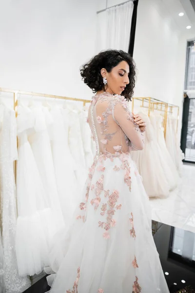Elegant middle eastern and brunette woman with wavy hair standing in gorgeous and floral wedding dress inside of luxurious bridal salon around white tulle fabrics, bridal shopping, looking away — Stock Photo