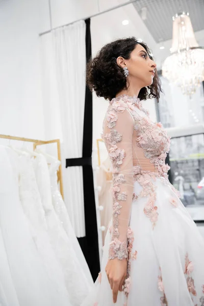 Bride-to-be, stunning middle eastern and brunette woman with wavy hair standing in gorgeous and floral wedding dress inside of luxurious bridal salon around white tulle fabrics, bridal shopping — Stock Photo