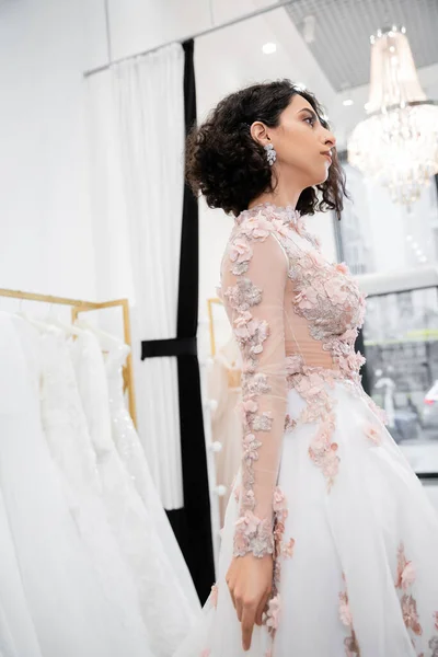 Novia-a-ser, impresionante mujer de Oriente Medio y morena con el pelo ondulado de pie en vestido de novia hermoso y floral dentro de lujoso salón nupcial alrededor de telas de tul blanco, compras nupciales - foto de stock