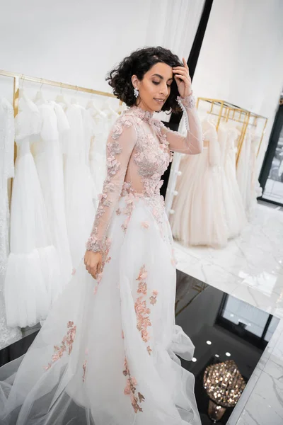 Bride-to-be, charming middle eastern and brunette woman with wavy hair standing in gorgeous and floral wedding dress inside of luxurious bridal salon around white tulle fabrics, shopping — Stock Photo
