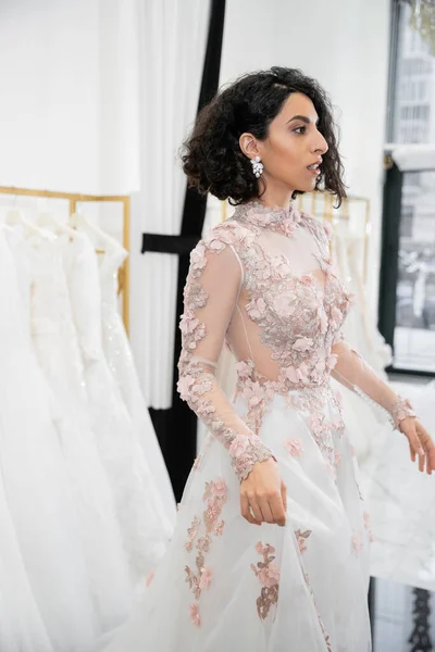 Bride-to-be, surprised middle eastern and brunette woman with wavy hair standing in floral wedding dress inside of luxurious bridal salon around white tulle fabrics, bridal shopping, looking away — Stock Photo