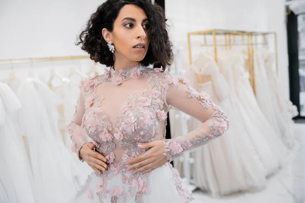 Stunning middle eastern and brunette bride with wavy hair standing in gorgeous and floral attire inside of luxurious bridal salon around white tulle fabrics, wedding dress shopping — Stock Photo