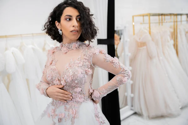 Bride-to-be, alluring middle eastern and brunette woman with wavy hair standing in gorgeous and floral wedding dress inside of luxurious salon around white tulle fabrics, bridal shopping — Stock Photo
