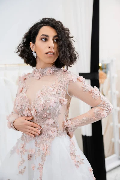Bride-to-be, brunette middle eastern woman with wavy hair standing in gorgeous and floral wedding dress inside of luxurious salon around white tulle fabrics, bridal shopping, sophisticated — Stock Photo