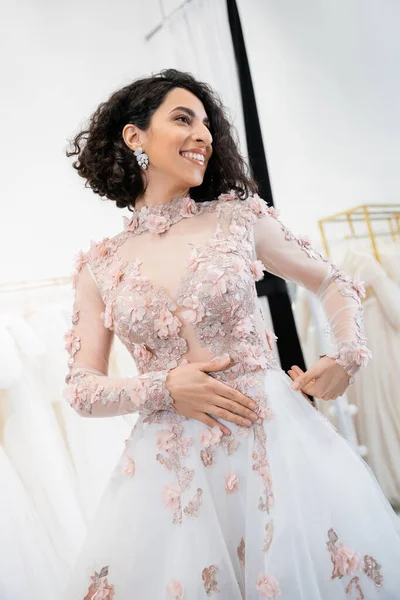 Enchanteresse Moyen-Orient et femme brune avec des cheveux ondulés debout dans une robe de mariée étonnante et florale et regardant loin à l'intérieur du salon de luxe autour de tissus en tulle blanc, achats nuptiaux — Photo de stock