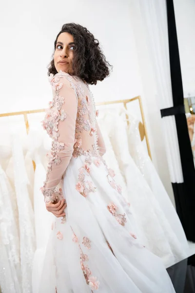 Entzückende Frau aus dem Mittleren Osten mit welligem Haar, die in einem wunderschönen und floralen Hochzeitskleid steht und von innen in einen luxuriösen Salon um weiße Tüll-Stoffe schaut, Brautshopping, zukünftige Braut — Stockfoto