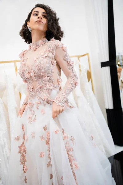 Attractive middle eastern woman with wavy hair standing in gorgeous and floral wedding dress and looking away inside of luxurious salon around white tulle fabrics, bridal shopping, bride-to-be — Stock Photo