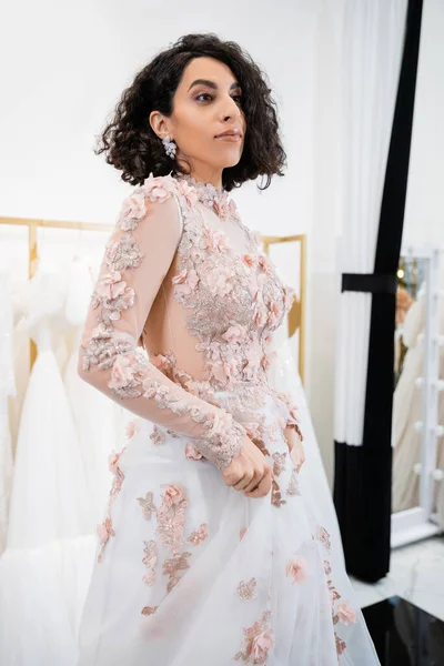 Завораживающая женщина с волнистыми волосами, стоящая в цветочном свадебном платье и смотрящая в роскошный салон вокруг белых тканей тюля, свадебные покупки, невеста-будущая — стоковое фото