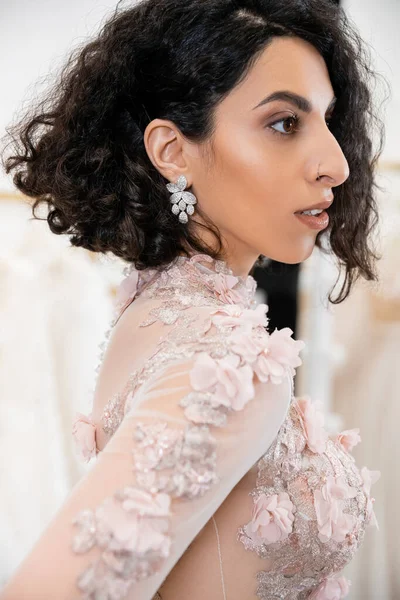 Portrait de charmante femme du Moyen-Orient avec des cheveux ondulés debout dans une robe de mariée magnifique et florale et regardant loin à l'intérieur du salon de luxe autour de tissus en tulle blanc, achats nuptiaux — Photo de stock