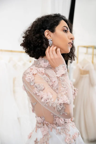 Delightful middle eastern woman with wavy hair standing in gorgeous and floral wedding dress and touching earring inside of luxurious salon around white tulle fabrics, bridal shopping — Stock Photo