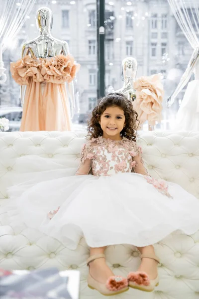 Cheerful middle eastern girl with curly hair sitting in floral dress on white couch inside of luxurious wedding salon, smiling kid, tulle skirt, bridal, blurred mannequin on background — Stock Photo