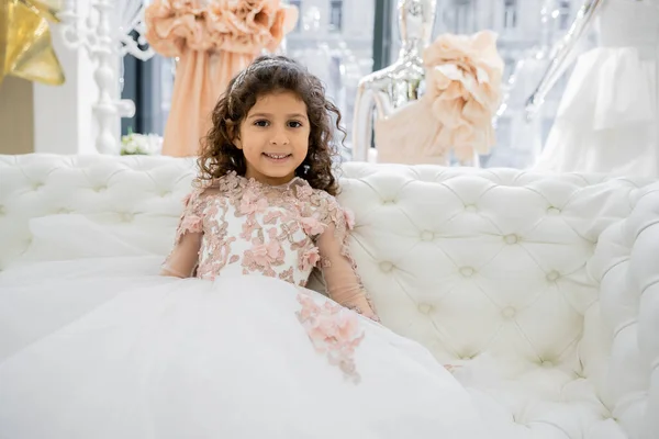 Веселая средневосточная девушка с вьющимися волосами, сидящая в цветочном платье на белом диване и улыбающаяся внутри роскошного свадебного салона, улыбающийся ребенок, юбка тюля, размытый фон — стоковое фото