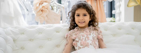 Glückliches mittelöstliches Mädchen mit lockigem Haar, das in einem floralen Kleid auf einer weißen Couch sitzt und in einem luxuriösen Hochzeitssalon lächelt, lächelndes Kind, Tüllrock, verschwommener Hintergrund, Banner — Stockfoto