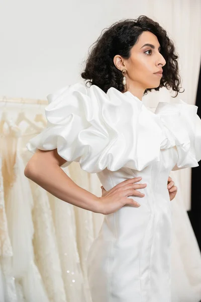 Brunette middle eastern bride with brunette and wavy hair posing with hands on hips in trendy wedding dress with puff sleeves and ruffles in bridal salon next to tulle fabrics — Stock Photo