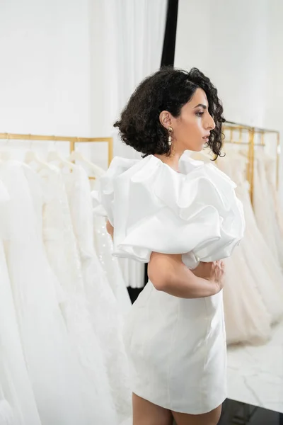 Brunette middle eastern bride with brunette and wavy hair posing in trendy wedding dress with puff sleeves and ruffles in bridal boutique next to tulle fabrics, white gown, side view — Stock Photo