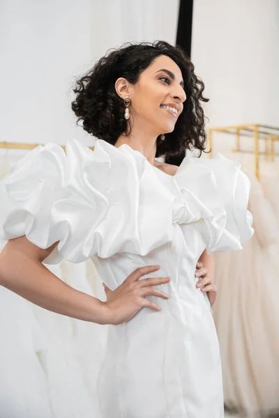 Cheerful middle eastern bride with brunette and wavy hair posing with hands on hips in trendy wedding dress with puff sleeves and ruffles in bridal salon next to tulle fabrics — Stock Photo