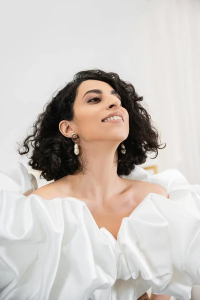 Portrait of happy middle eastern bride with brunette and wavy hair posing in trendy wedding dress with puff sleeves and ruffles looking up in bridal salon, white gown — Stock Photo
