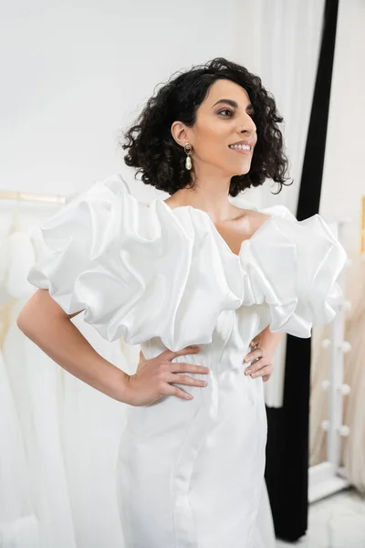 Positive middle eastern woman with brunette and wavy hair posing with hands on hips in trendy wedding dress with puff sleeves and ruffles in bridal salon next to tulle fabrics — Stock Photo