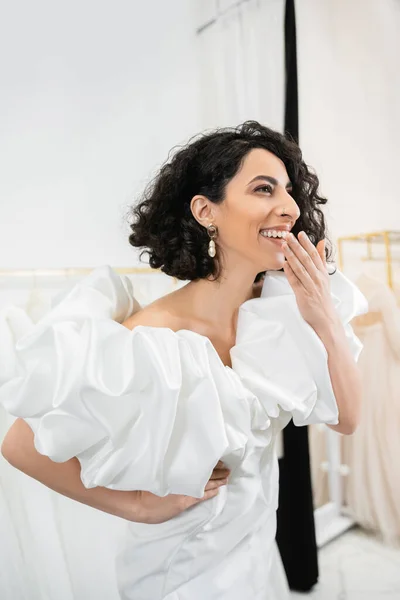 Radiant middle eastern bride with brunette wavy hair laughing, covering mouth and posing in trendy wedding dress with puff sleeves and ruffles in bridal salon, fashion — Stock Photo