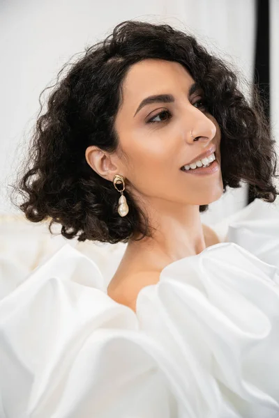 Portrait of happy brunette middle eastern bride with wavy hair posing in trendy wedding dress with puff sleeves and ruffles in bridal boutique and looking away, delightful woman — Stock Photo