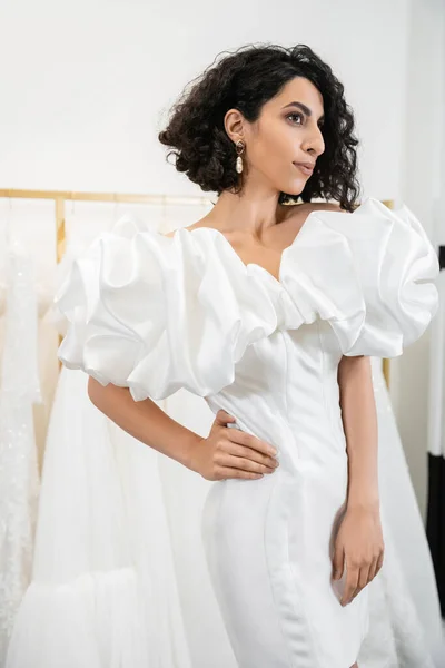 Brunette middle eastern bride with brunette and wavy hair posing with hand on hip in stylish wedding dress with puff sleeves and ruffles in bridal boutique next to tulle fabrics, delightful woman — Stock Photo