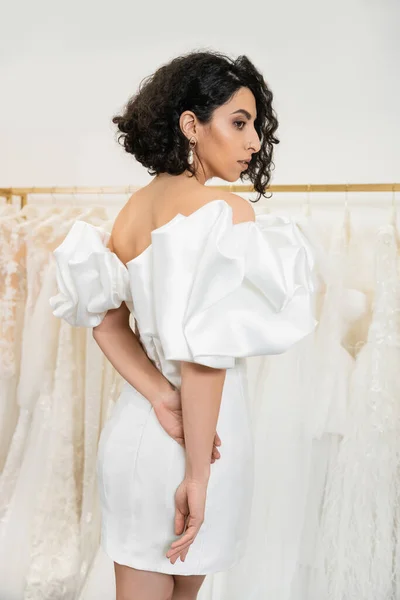 Stunning middle eastern bride with brunette and wavy hair posing in stylish wedding dress with puff sleeves and ruffles in bridal boutique next to tulle fabrics, elegant woman — Stock Photo