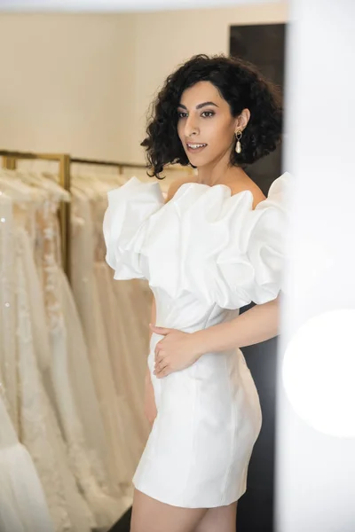Smiling middle eastern woman with brunette and wavy hair trying on trendy wedding dress with puff sleeves and ruffles near mirror in bridal boutique next to tulle fabrics, reflection, shopping — Stock Photo