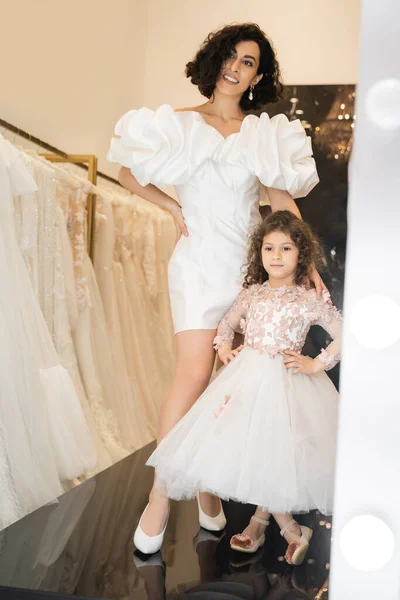 Joyful middle eastern bride with wavy hair posing in trendy wedding dress with puff sleeves and ruffles near cute little daughter in floral attire and white bridal gown in boutique — Stock Photo