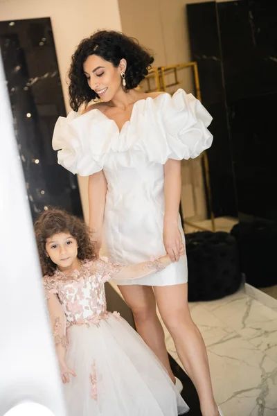 Happy middle eastern bride with wavy hair standing in trendy wedding dress with puff sleeves and ruffles while looking at cute little daughter in floral attire in bridal boutique — Stock Photo